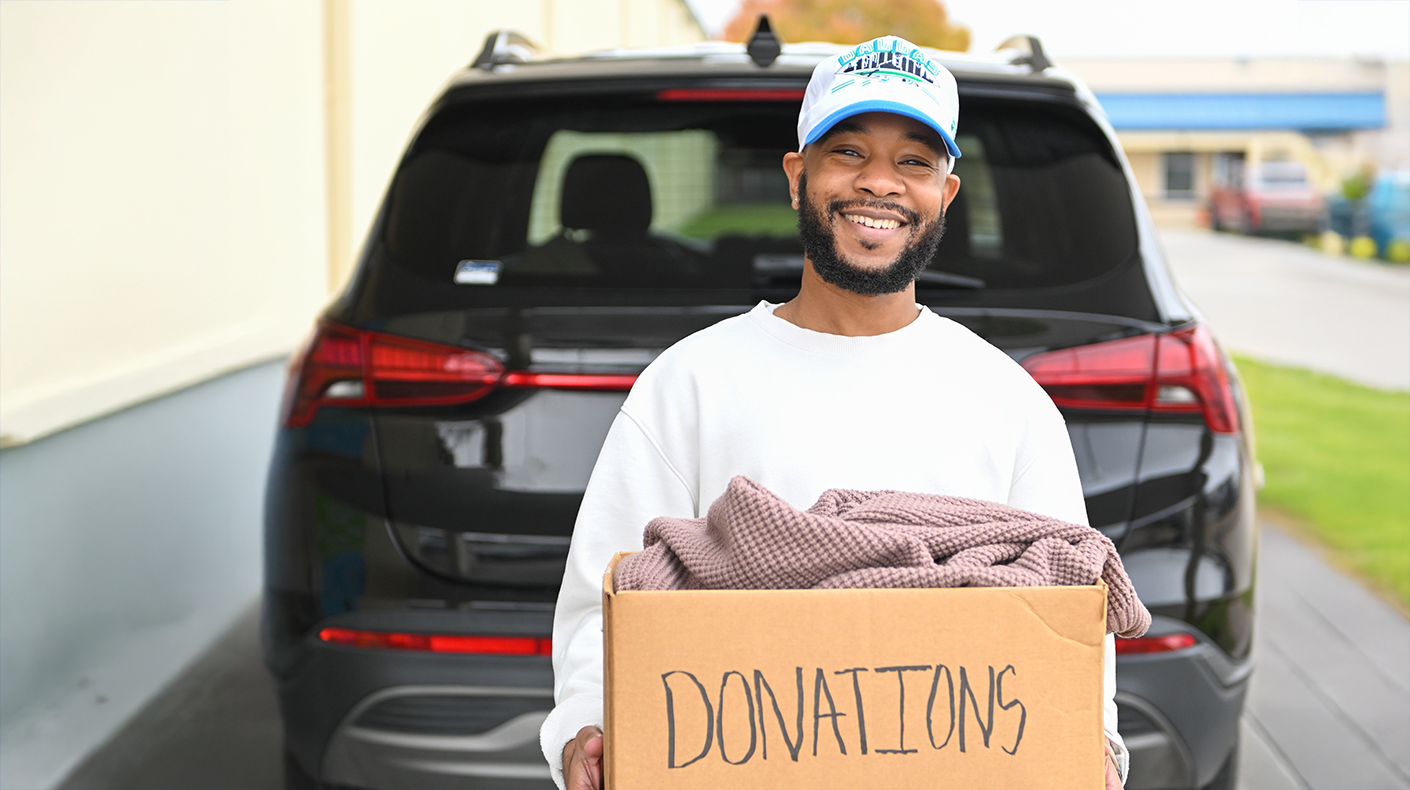 Man holding a box labeled donations posing in front of open car trunk.
